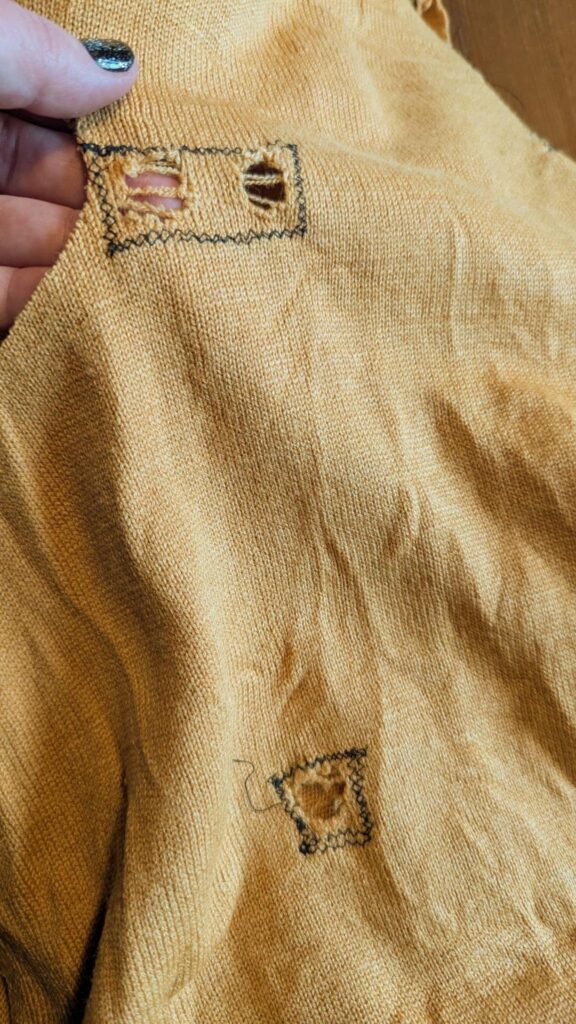 Close photo of my hand holding a piece of mustard yellow, woolly knit fabric with holes that have been sewn around in black thread to stop them from running. I have pale skin and a black manicure with gold sparkles chosen by a child