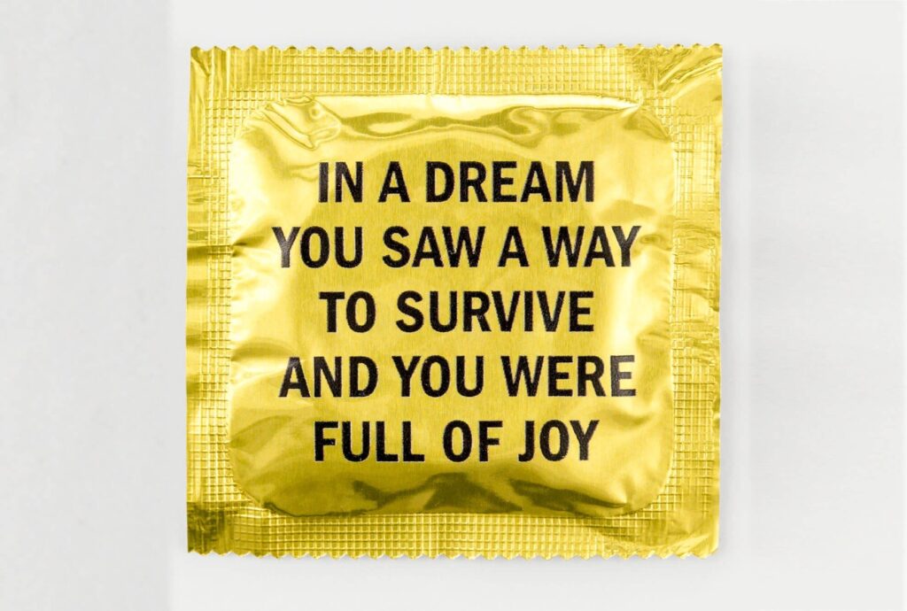 A condom package made of gold foil, with black text printed on it that reads "IN A DREAM YOU SAW A WAY TO SURVIVE AND YOU WERE FULL OF JOY"