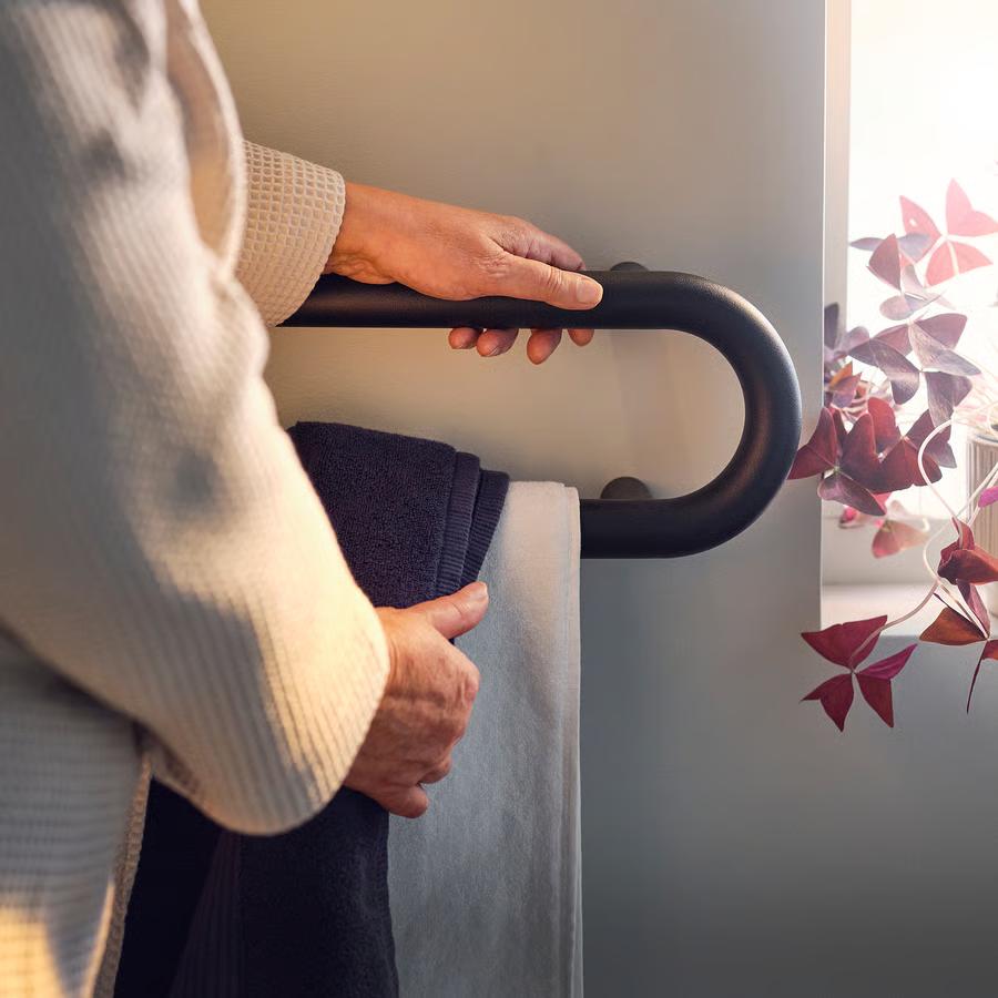 Catalog photo of a person in a white bathrobe pulling a towel off a rack while resting the other hand on the towel rack to stabilize. The rack is a black tube looped into a pill shape, with four anchor points.