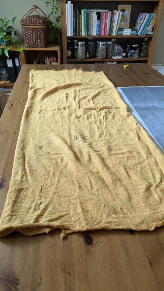 Photo of a wooden table on which is a ratty piece of mustard yellow fabric and a pattern made from 6 pieces of printer paper taped together. In the background are shelves with jars, books, plants, etc