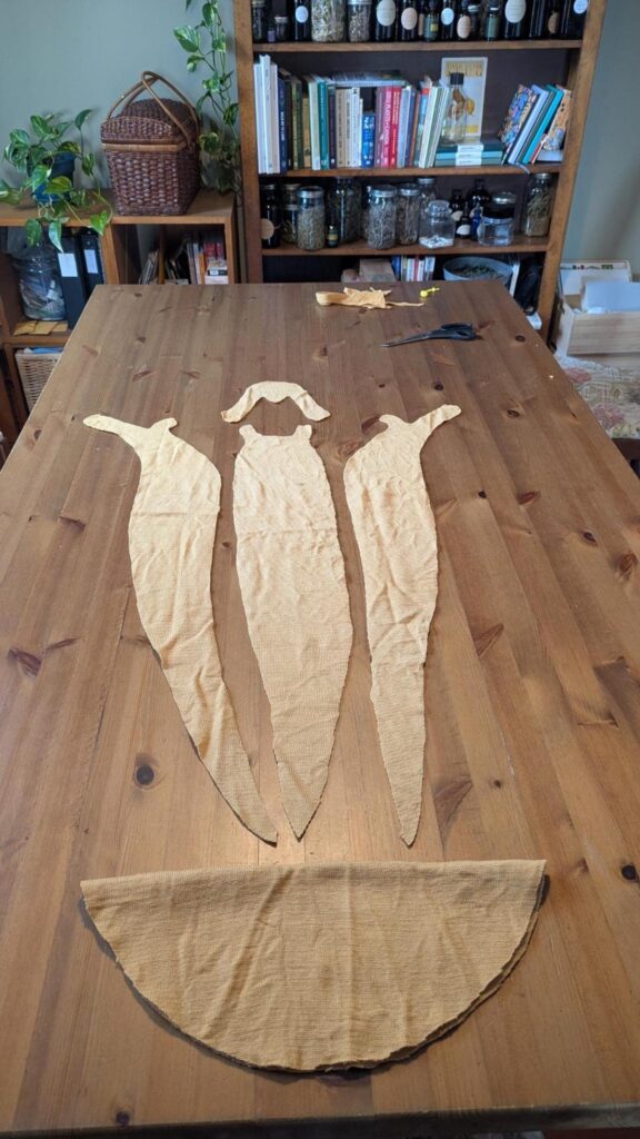 Photo of a wooden table on which are laid out flat pieces of mustard yellow fabric: 3 long slug body shapes, a horned slug face piece, and a folded half-circular slug mantle