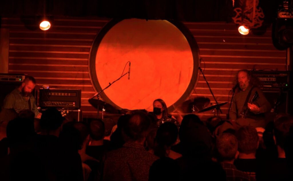 Dark photo of a nightclub stage where three musicians are playing under red lighting. In the center, the drummer is wearing a black, ear-loop mask.