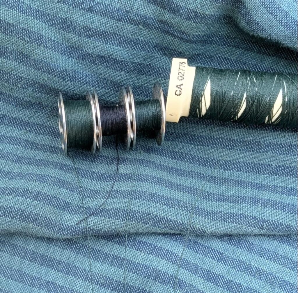 Close photo of dark teal and blue striped fabric with different threads laid out on it. The first two bobbins are a dark green and a navy blue, and their trailing threads contrast against the fabric. Next are a bobbin and a partly-used spool of the same dark bluish-green. Those trailing threads blend into the background fabric and are hard to see.