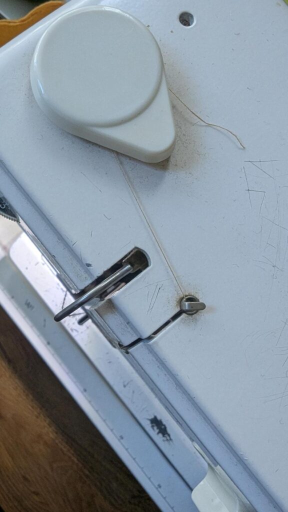 Photo of the top of a sewing machine, showing a tiny tail of tan thread sticking out from that first doohickey that the thread goes around