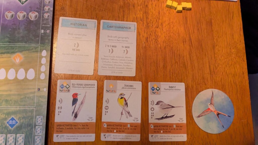 Photo of a Wingspan board game in progress. Three cards have illustrations of birds with the names red-headed woodpecker, dickcissel, and bushtit
