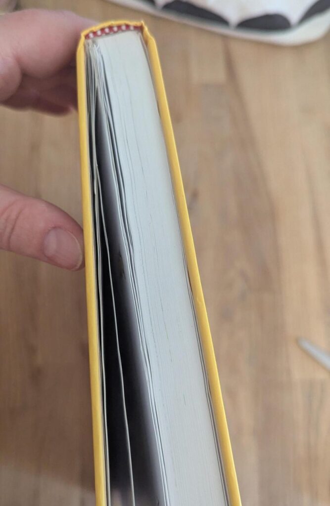 Photo of my pale hand holding a closed handcover book, with the top edge visible to show a section of lumpy and uneven pages where they have been taped back in unevenly
