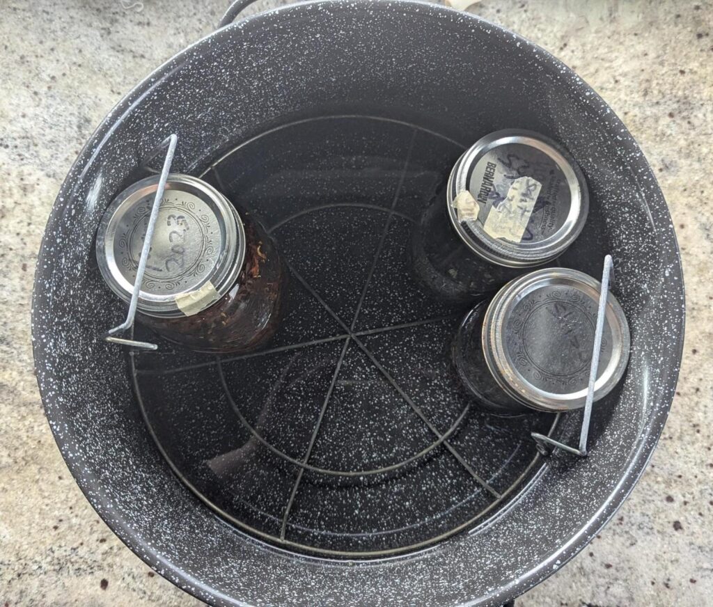 Photo looking down into a speckled black canning pot containing a wire rack and 3 mason jars with old labels on the lids.